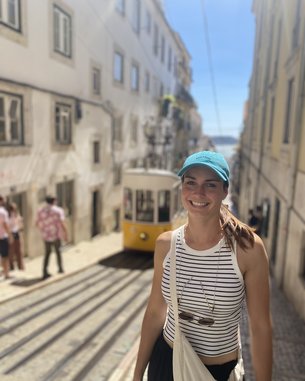 Eine Frau mit einem blauen Cap und einem gestreiften Oberteil steht in einer engen Straße, während eine gelbe Straßenbahn vorbeifährt. Im Hintergrund sind weitere Personen und historische Gebäude sichtbar.