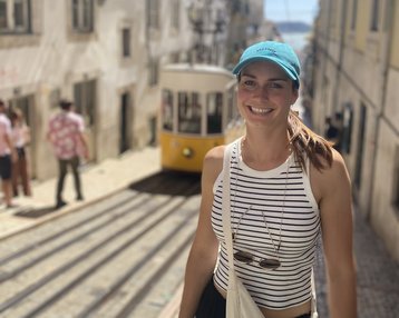 Eine Frau mit einem blauen Cap und einem gestreiften Oberteil steht in einer engen Straße, während eine gelbe Straßenbahn vorbeifährt. Im Hintergrund sind weitere Personen und historische Gebäude sichtbar.