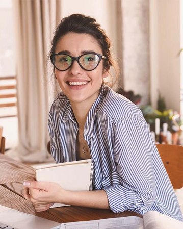 Eine lächelnde Frau mit Brille sitzt an einem Tisch, umgeben von Pflanzen und Blumen, während sie ein Notizbuch hält. Ein Laptop steht vor ihr.