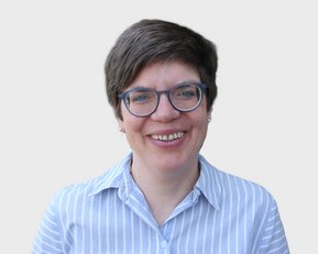 Eine lächelnde Person mit kurzen, grauen Haaren und einer blauen, gestreiften Bluse trägt eine Brille.