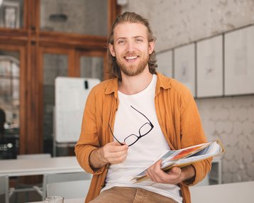 Ein junger Mann mit langen Haaren sitzt auf einem Tisch, hält eine Mappe mit Unterlagen und eine Brille in der Hand. Er trägt ein orangefarbenes Hemd und lächelt.