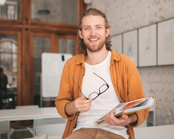 Ein junger Mann mit langen Haaren sitzt auf einem Tisch, hält eine Mappe mit Unterlagen und eine Brille in der Hand. Er trägt ein orangefarbenes Hemd und lächelt.