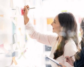 Zwei Frauen arbeiten gemeinsam an einer Wand mit Haftnotizen. Eine Frau schreibt mit einem Stift auf eine Haftnotiz, während die andere sie beobachtet und Notizen macht.