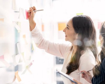 Zwei Frauen arbeiten gemeinsam an einer Wand mit Haftnotizen. Eine Frau schreibt mit einem Stift auf eine Haftnotiz, während die andere sie beobachtet und Notizen macht.