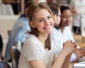 Eine lächelnde Frau mit langen, blonden Haaren sitzt an einem Tisch, umgeben von anderen Personen in einem modernen Büro.