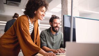 Eine Frau mit lockigem Haar und einem braunen Mantel steht neben einem Mann mit Bart, der an einem Computer arbeitet. Beide lächeln und scheinen an einem Projekt interessiert zu sein.