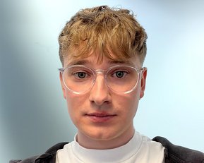 Junger Mann mit kurzen, lockigen Haaren und Brille, trägt ein weißes T-Shirt und einen schwarzen Hoodie, schaut direkt in die Kamera.