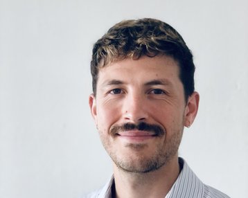 Ein Mann mit einem Schnurrbart trägt ein gestreiftes Hemd und lächelt in die Kamera. Der Hintergrund ist hell und neutral.