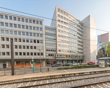 Moderne Bürogebäude mit einer markanten, schrägen Fassade, umgeben von Straßen und Gleisen. Im Vordergrund sind Personen und eine Haltestelle zu sehen.