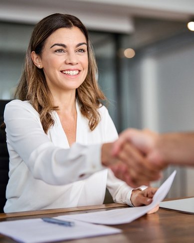 Eine Frau in einem weißen Hemd schüttelt die Hand eines Mannes am Tisch. Auf dem Tisch liegen einige Dokumente und ein Laptop.