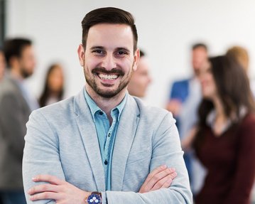 Ein Mann im Anzug mit einem breiten Lächeln und gekreuzten Armen steht im Vordergrund, während eine Gruppe von Menschen im Hintergrund miteinander spricht.