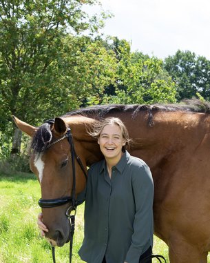 Eine Person steht neben einem braunen Pferd und lächelt. Die Person trägt ein graues Hemd und hält das Pferd am Halfter. Im Hintergrund sind Bäume und eine grüne Wiese zu sehen.
