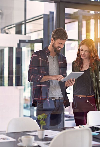 Zwei Personen stehen in einem modernen Büro und betrachten gemeinsam ein Tablet. Der Mann trägt ein kariertes Hemd, die Frau eine Jacke. Auf dem Tisch sind Unterlagen und Tassen sichtbar.