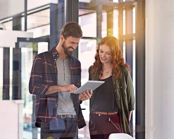 Psychologie in Organisationen | SRH Fernhochschule Zwei Personen stehen in einem modernen Büro und betrachten gemeinsam ein Tablet. Der Mann trägt ein kariertes Hemd, die Frau eine Jacke. Auf dem Tisch sind Unterlagen und Tassen sichtbar.