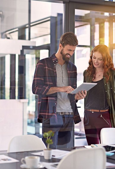 Zwei Personen stehen in einem modernen Büro und betrachten gemeinsam ein Tablet. Der Mann trägt ein kariertes Hemd, die Frau eine Jacke. Auf dem Tisch sind Unterlagen und Tassen sichtbar.