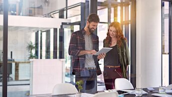 Psychologie in Organisationen | SRH Fernhochschule Zwei Personen stehen in einem modernen Büro und betrachten gemeinsam ein Tablet. Der Mann trägt ein kariertes Hemd, die Frau eine Jacke. Auf dem Tisch sind Unterlagen und Tassen sichtbar.