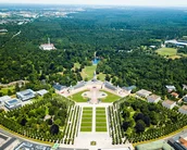 Luftaufnahme eines großen Parks mit symmetrischen Gartenanlagen, Wegen und einem zentralen Brunnen. Umgeben von Bäumen und modernen Gebäuden, erstreckt sich der Park in eine grüne Landschaft.