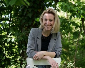 Eine Frau sitzt lässig auf einem weißen Hocker, trägt eine graue Jacke und Jeans, umgeben von grünen Pflanzen.