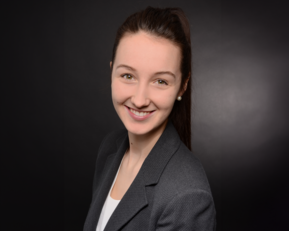 Eine junge Frau mit einem freundlichen Lächeln trägt einen grauen Blazer und hat lange, dunkle Haare. Der Hintergrund ist dunkel.