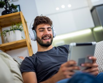 Junger Mann mit Kopfhörern, der auf einem Tablet sitzt und lächelt, während er entspannt auf einem Sofa ist.