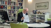 Ein Mann in einem Anzug sitzt an einem Tisch und liest Dokumente. Im Hintergrund sind Bücherregale und ein Computer sichtbar.