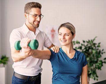 Ergotherapie Bachhelor-Studium an der SRH Fernhochschule In einer Ergotherapie Sitzung hilft ein Mann einer Frau beim Heben einer Hantel während einer Übungssitzung. Die Frau lächelt, während der Mann sie an der Schulter unterstützt.
