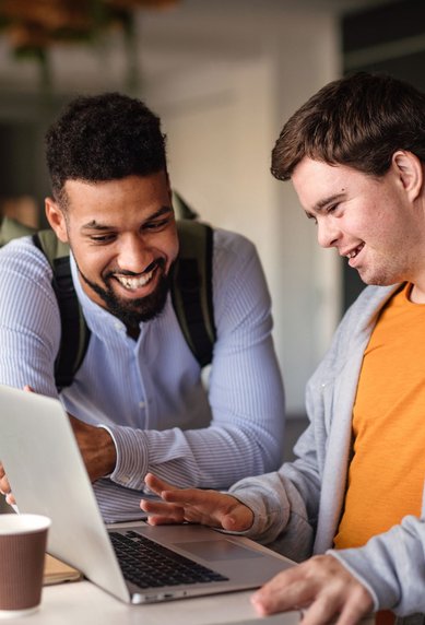 Zwei Männer stehen an einem Tisch und schauen auf einen Laptop. Der Mann links lächelt und zeigt auf den Bildschirm, während der Mann rechts interessiert zuschaut. Eine Kaffeetasse steht auf dem Tisch.