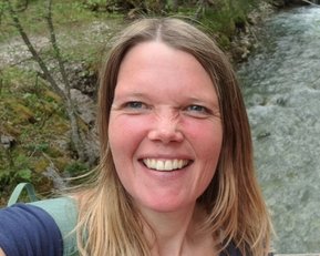 Eine Frau mit langen, blonden Haaren lächelt in die Kamera, während sie an einem Flussufer steht, umgeben von Bäumen.