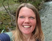 Eine Frau mit langen, blonden Haaren lächelt in die Kamera, während sie an einem Flussufer steht, umgeben von Bäumen.