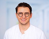 Ein lächelnder Arzt trägt eine weiße Kittel und runde Brille. Der Hintergrund ist unscharf und hell, was den Fokus auf die Person lenkt.