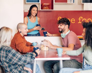 Eine Gruppe junger Menschen sitzt entspannt an einem Tisch und unterhält sich bei Kaffee oder Tee. Eine weitere Person steht lächelnd hinter einer Theke mit der Aufschrift „free food“. Die Atmosphäre wirkt freundlich und einladend, wie in einem Gemeinschaftsraum oder sozialen Treffpunkt.