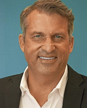 Ein Mann mit kurzen, grauen Haaren und einem freundlichen Lächeln trägt einen schwarzen Anzug und ein weißes Hemd.