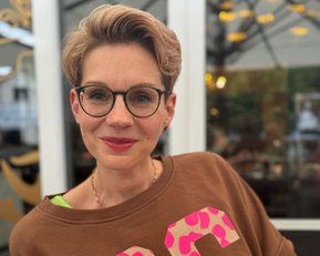 Frau mit kurzen, blonden Haaren und Brille trägt einen braunen Pullover mit großen, pinken Zahlen. Sie lächelt und steht vor einem hellen Hintergrund mit unscharfen Details.