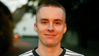 Junger Mann mit kurzen Haaren trägt ein gestreiftes T-Shirt und lächelt in die Kamera, während er auf einem Weg steht.