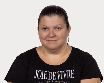 Eine Frau mit kurzen, dunklen Haaren trägt ein schwarzes T-Shirt mit dem Aufdruck „JOIE DE VIVRE“ und schaut direkt in die Kamera.