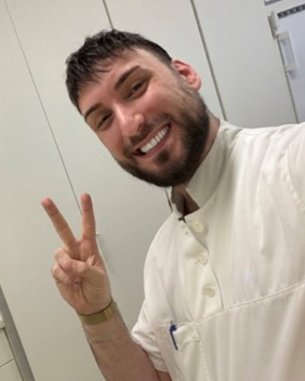 Ein Mann mit Bart und lächelndem Gesicht trägt eine weiße Uniform und zeigt ein Peace-Zeichen mit der Hand.