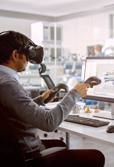 Ein Mann trägt ein Virtual-Reality-Headset und hält zwei Controller in einem modernen Büro. Auf dem Bildschirm vor ihm ist ein 3D-Modell zu sehen. Im Hintergrund arbeitet eine weitere Person an einem Computer.