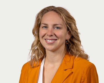 Eine lächelnde Frau mit lockigem, blondem Haar trägt ein orangefarbenes Blazer. Der Hintergrund ist neutral und hell.