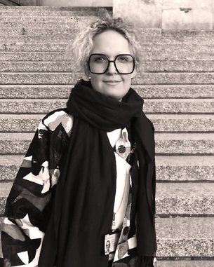 Eine Person mit lockigem Haar und großen Brillen steht auf einer Treppe. Sie trägt ein auffälliges, gemustertes Oberteil und einen schwarzen Schal.