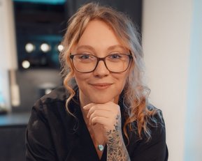 Eine Person mit Brille und tätowiertem Arm lächelt, während sie auf einem Tisch in einer modernen Küche sitzt.