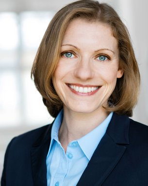 SRH Fernhochschule | Alumni-Portrait des Monats Eine lächelnde Frau mit blonden Haaren trägt einen dunklen Blazer und ein hellblaues Hemd. Der Hintergrund ist unscharf und hell, was den Fokus auf ihr Gesicht lenkt.