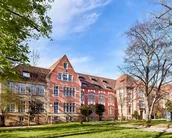 Historische Gebäude mit roten Ziegeln und großen Fenstern, umgeben von Rasenflächen und Bäumen. Der Himmel ist klar und blau, was eine sonnige Atmosphäre vermittelt.