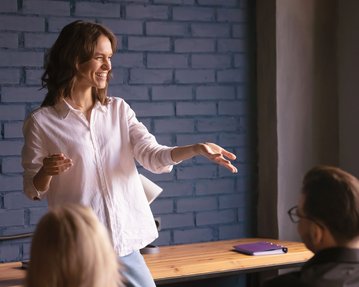 Eine Frau in einem weißen Hemd spricht und gestikuliert vor einem Publikum, das ihr aufmerksam zuhört. Im Hintergrund sind eine blaue Wand und ein Tisch mit Notizmaterialien sichtbar.
