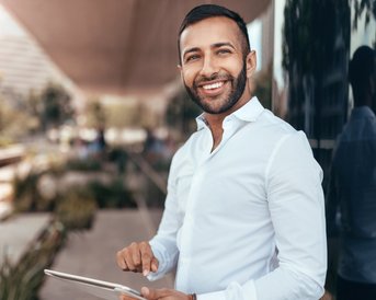 Ein Mann in einem weißen Hemd steht draußen und hält ein Tablet in der Hand. Er lächelt und blickt in die Kamera, während er vor einer modernen Gebäudeoberfläche steht.