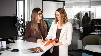 Zwei Frauen stehen an einem Schreibtisch in einem modernen Büro und besprechen Notizen. Eine Frau hält einen Stift und schreibt in ein Notizbuch.