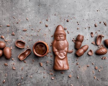 Ein Schokoladenfigur eines Weihnachtsmanns umgeben von Schokoladeneiern und einer kleinen Schüssel mit Schokolade auf einem grauen Untergrund.