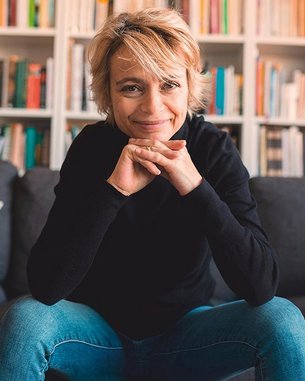 Eine Person mit blonden, kurzen Haaren sitzt auf einem Sofa, lächelt und stützt ihr Kinn auf die Hände. Im Hintergrund sind Bücherregale mit zahlreichen Büchern sichtbar.