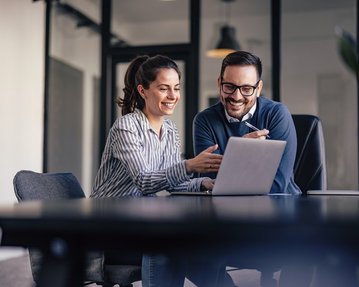 Krisenkommunikation in Unternehmen Zwei Personen sitzen an einem Tisch und schauen auf einen Laptop. Die Frau zeigt auf den Bildschirm, während der Mann lächelt und aufmerksam zuhört. Beide scheinen an einer Diskussion oder Präsentation beteiligt zu sein.