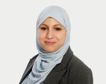 Eine Frau mit einem hellblauen Hijab und einem grauen Blazer lächelt in die Kamera. Der Hintergrund ist neutral und hell.