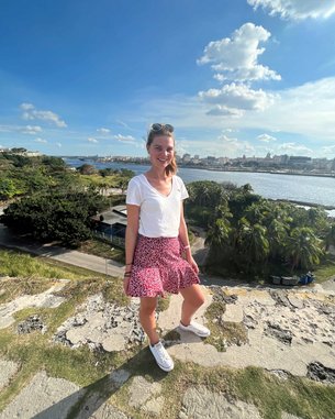 Eine junge Frau steht auf einer Mauer mit Blick auf eine Stadtlandschaft und einen Fluss. Sie trägt ein weißes T-Shirt, einen gemusterten Rock und Sneakers. Im Hintergrund sind Bäume und Gebäude sichtbar.